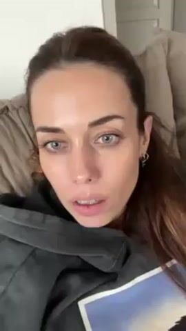 A woman with long brown hair pulled back in a partial ponytail is lying back on a tan, cushioned couch, presumably indoors. She is wearing a gray hoodie with a blue image on it and small hoop earrings. She is looking directly at the camera, opening and closing her eyes, and talking. She says her eyes are red from spring allergies. The background includes a door and part of a light-colored wall.