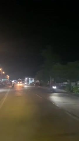 The video captures a nighttime view from a moving vehicle on a road, likely in a semi-urban or rural area. The road is paved and illuminated by streetlights and vehicle headlights. Several motorcycles and a truck are visible, driving in both directions. Buildings, some with lit windows, line the side of the road, suggesting a commercial or residential area. Trees are present on the right side of the road. The overall atmosphere is that of a typical night scene on a road with active traffic.