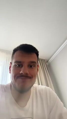 A man with short dark hair, a mustache, and wearing a white t-shirt is speaking directly to the camera. He is indoors, with white walls and ceiling visible behind him, along with beige curtains. At the beginning of the video, he looks up briefly and raises his index finger. He then brings his hand to his face to cover his mouth.