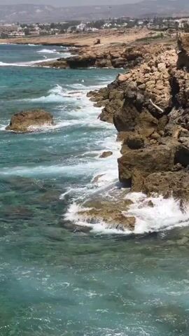 This is a medium shot video of a rocky coastline with clear turquoise water and waves crashing against the rocks. In the background, there is a town or village with buildings and some greenery. The sky is bright and hazy. The camera is stationary.