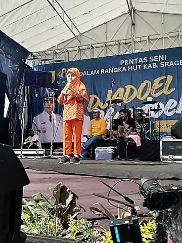 A young girl stands on a stage speaking into a microphone. She is wearing an orange patterned outfit and a hijab. Behind her, there is a banner for "Pentas Seni Dalam Rangka HUT Kab. Sragen KE-278" with "Djadoel Djadoel" written in large font. On the stage are three seated men and a drum set. The stage is set up under a white tent. In the foreground, there is a professional camera on a tripod, suggesting the event is being recorded.