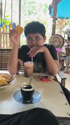 A young boy sits at a table with food and drinks. He holds a piece of fried food in his right hand, looks at the camera, and stirs his chocolate drink with a straw with his left hand. On the table, there is a plate with fried food, a glass of black coffee, a glass of chocolate drink, and a package of crackers. The table is white and has some stains on it.  The boy wears a black t-shirt with a white square containing a "U" design on it. He has black hair and a serious expression. Behind him is a wooden bench, a window with greenery visible, and some tools or equipment hanging from the ceiling. The setting looks like an outdoor seating area.