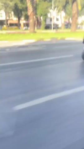 The video shows a street scene with palm trees lining a median. Cars are driving on the road. In the background, there are buildings, some of which appear to be businesses. The sky is blue and there are some clouds. The video is shot from a moving vehicle.