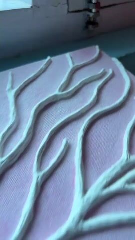 The video shows a close-up of a pink canvas with a white sculpted tree branch design. Small white flowers are attached to the branches. The canvas is placed on a surface with a striped pattern, and the background shows a window with plants on the windowsill. The camera slowly pans out, revealing the entire canvas and its placement.