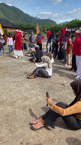 A group of people are gathered outdoors, seemingly for a game or event. In the foreground, two women are seated on the ground. One woman, wearing a brown hijab and a brown long-sleeved shirt, is looking towards a group of people further away and gesturing with her left hand. She is wearing light blue wide-leg pants and dark sandals. To her right, another woman wearing a hijab and an orange long-sleeved shirt is also seated and holding a dark object, possibly a phone. She is wearing striped pants and sandals. In the mid-ground, several people are sitting or kneeling on the ground, some appearing to be participants in a game. One person in a white and black hijab is in the center, holding their hands up. A woman in a red outfit and a white hijab is standing to the left and appears to be leading or instructing. Other people, including children, are standing and watching the scene. There are flags, including a yellow and white flag, and red and white flags, visible in the background. The environment appears to be a sandy or gravelly area with hills and vegetation in the distance, under a partly cloudy sky. Bottles are visible on the ground near the seated participants.