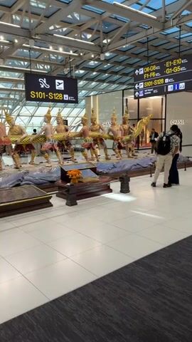 The video shows an airport interior with a large, ornate sculpture of figures pulling a serpent-like creature. The sculpture is positioned in the middle of a wide, tiled walkway. Several people are visible in the background, walking or standing. One couple in the foreground is looking at the sculpture, with the man wearing a backpack and the woman in a white top and black pants. Signage is visible in the background, including gate information and airline logos. The environment appears to be a modern airport terminal with a high, glass ceiling. The sculpture itself is detailed, with figures in traditional attire and the serpent depicted with scales. A sign in Thai and English reading "PLEASE DO NOT TOUCH" is placed near the base of the sculpture.