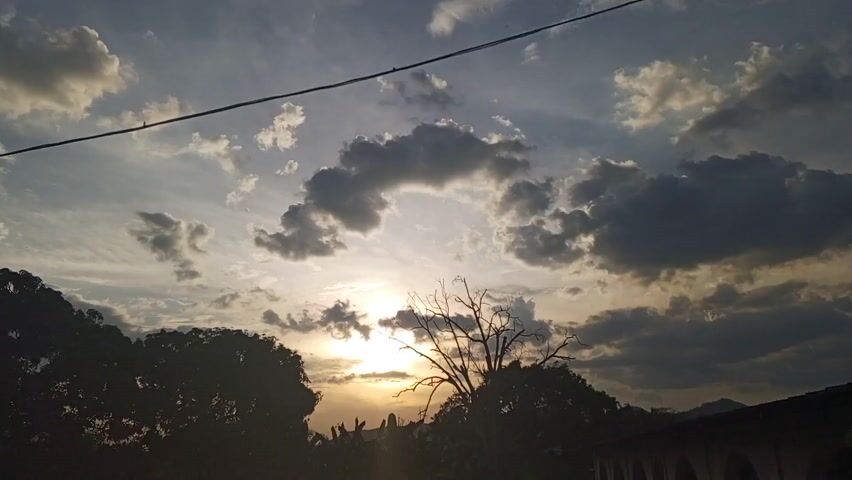 The video shows a sunset sky with clouds. The sun is visible through the clouds, and there are trees silhouetted against the sky in the foreground. A power line is visible in the upper part of the frame. There is a mountain range barely visible in the distance.