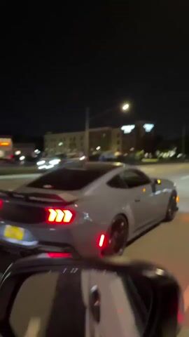 The video captures a gray Ford Mustang GT (S650 generation) accelerating loudly on a street at night. The car is seen from the rear, slightly to the right, with its distinctive triple-bar red taillights illuminated. As the video begins, the Mustang is stationary or moving slowly, then it rapidly accelerates, producing a loud, aggressive exhaust note with audible pops and crackles.

The scene is set on a multi-lane road, likely in an urban or suburban area, consistent with the Alexandria, USA context. In the background, streetlights illuminate the road, and residential or commercial buildings are visible further down the street. The sky is dark, indicating it's nighttime. The road appears dry, and visibility is good, illuminated by artificial light sources.

A yellow license plate with "SUDDY 7" is clearly visible on the rear of the Mustang. The recording device is inside another vehicle, with a portion of its interior and dashboard visible in the lower-left corner of the frame. The event portrays a display of the Mustang's performance capabilities, specifically its acceleration and exhaust sound.