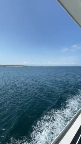 The video captures a wide, serene view of a large body of water, likely the sea or ocean, under a bright blue sky dotted with scattered white clouds. The perspective is from a moving boat, indicated by the foamy white wake trailing in the bottom right corner and the partial view of the boat's white and dark grey structure along the right edge of the frame. In the distance, a low-lying, sandy-brown coastline stretches across the horizon, suggesting a landmass or island. The water is a deep blue-green hue with gentle ripples. The scene is brightly lit, indicating a clear, sunny day.