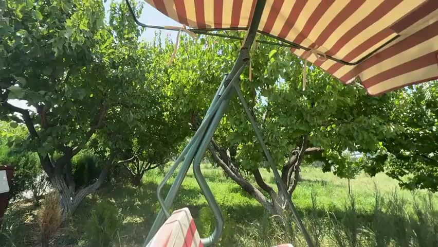 The video shows a swing set with a striped awning in what appears to be a backyard or park setting. Green trees and foliage are visible in the background, along with a small wooden structure that might be a playhouse. There is also a seesaw with red handles. The sky is blue and sunny, suggesting it is daytime. The camera pans around the scene, offering different views of the swing set and its surroundings. There are no people visible in the video.