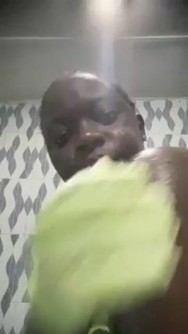 A person with dark skin, facing the camera, is in a shower. They are using a light green exfoliating glove to scrub their shoulder and upper arm, which is covered in a white, soapy substance. The background features white tiles with a light blue geometric pattern. The lighting in the shower is bright.  The person smiles briefly at the camera.