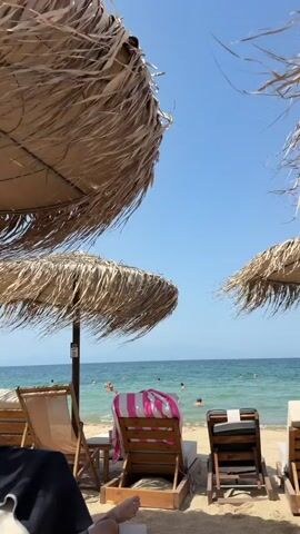 The video shows a view of a beach from under two straw umbrellas. In the foreground, there are several empty lounge chairs. The sea is visible in the background with several people swimming. The sky is clear and blue.