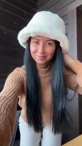 A woman with long, dark hair stands facing the camera, wearing a beige turtleneck sweater, light-colored pants, and a white fluffy hat. She blows a kiss to the camera, then touches her hair. The background is a wall of dark horizontal wood planks.