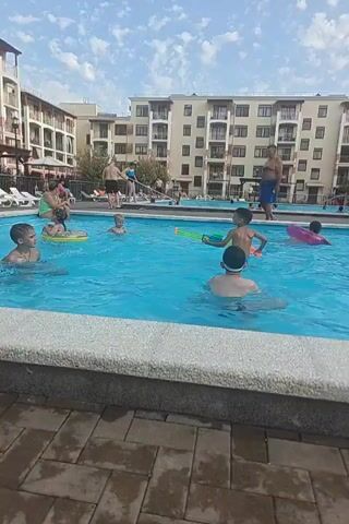 The video shows a busy outdoor swimming pool area. There are several people in the pool, mostly children, engaged in various activities. One child in the foreground is wearing swimming goggles. Another child is playing with a green toy. A woman in a dark swimsuit is in the middle of the pool, and a child on an inflatable pink ring is further back. Adults are also present, some standing by the pool and others in the water. In the background, there are multi-story buildings, likely apartments or hotel rooms, with balconies overlooking the pool. Lounge chairs are set up around the pool, and some people are relaxing on them. The sky is partly cloudy. The overall atmosphere suggests a lively and enjoyable summer day at a resort or hotel.