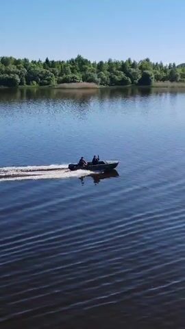 A motorboat carrying two individuals navigates a wide, calm body of water under a clear blue sky. The boat is moving from left to right across the frame, leaving a white wake behind it. The sun glints off the water's surface, creating sparkling reflections, particularly on the right side of the frame. In the background, a lush green treeline borders the far shore of the water body. The overall impression is one of peaceful movement on a bright, sunny day in what appears to be a natural, possibly rural, setting in Kargulino, Russia. No visible text is present.