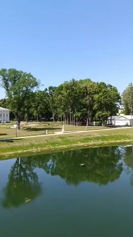 The video shows a serene park landscape with a large body of dark water in the foreground. The water reflects the lush green trees and the clear blue sky with scattered white clouds. On the far bank, a white building with a pitched roof is visible amongst the trees. Further to the right, a white gazebo with a domed roof stands prominently. In the distance, a white bridge with an arched structure spans across the water, with decorative oval shapes along its railing. The overall impression is of a tranquil, well-maintained park on a sunny day.