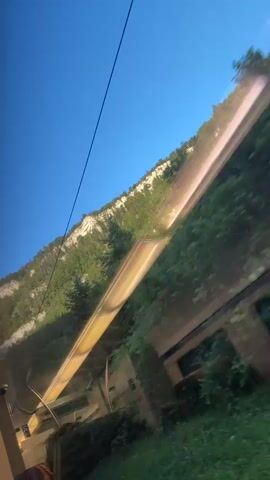 A person is looking out of a train window at a bright blue sky and a lush green mountainside. The train appears to be moving, as the scenery is blurred. A portion of the train's interior is visible in the foreground, including what looks like a yellow railing and part of a seat. The train is passing through a wooded area, with trees and foliage obscuring the base of the mountain. There are also some indistinct structures visible, which could be buildings or ruins, partially overgrown with vegetation.