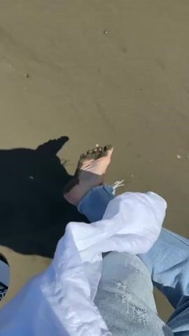 The video begins with a close-up shot of a person's feet on a sandy beach. The feet are bare and covered in dark wet sand. The person is wearing ripped blue jeans. A white garment is draped over their lap. The scene transitions to a wider shot of the beach and ocean. The sand is damp and shows patterns from receding waves. The ocean is calm with gentle waves and a clear blue sky with a few white clouds. In the distance, there are a couple of sailboats and a few people wading in the shallow water.