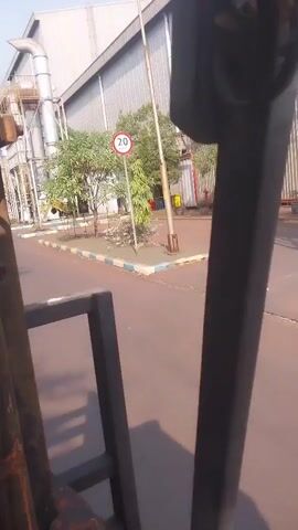 The video begins with a close-up view of a forklift's metal structure. The camera then pans to reveal an industrial setting with a long, corrugated metal building in the background. A speed limit sign indicating 20 km/h is visible. Trees and some debris are scattered around the area. The camera continues to pan, showing a road with yellow and black striped bollards lining the side. Further down the road, there are more industrial buildings and structures, including large pipes and tanks. The video ends with a shot of the road stretching into the distance, with several plastic containers visible at the bottom of the frame.