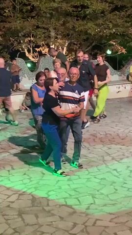 A group of people are dancing in an outdoor area with a mosaic tile floor and trees in the background. The lighting is dim, with some areas illuminated by green and purple lights. Several couples are dancing, and some people are standing around watching. In the foreground, a woman in a blue top and patterned pants is standing still, while a couple dances beside her. Further in the background, other people are dancing, and some are seated. The scene appears to be a social gathering or event.