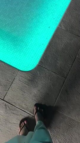 The video shows a person's feet at the edge of a swimming pool. The pool is illuminated with blue and green lights, and the water appears clear. The ground around the pool is paved with stone tiles. The video pans across the pool, showing its depth and tiled bottom. The scene appears to be at night, as suggested by the dark background and artificial lighting.