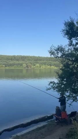 Two people are fishing from the shore of a calm lake. A man stands and casts his fishing rod while a child sits in a chair nearby. A small white boat with at least two people aboard is visible further out on the lake. The lake is surrounded by a dense, green forest, and the sky is clear and blue.