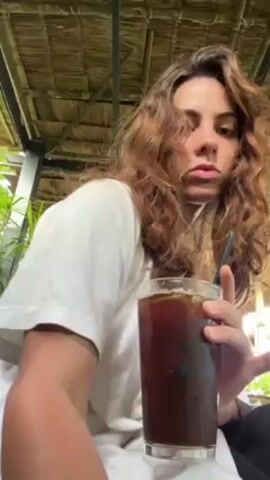 A young woman with long, curly brown hair sits outdoors under a thatched roof. She is wearing a white t-shirt and dark pants.  She holds a tall glass of dark liquid with ice, which appears to be iced coffee, in her left hand. With her right hand, she pours a small amount of white liquid, presumably milk or cream, from a small, clear glass container into the coffee. The coffee begins to mix with the added liquid, creating a swirling pattern. The drink sits on her lap.  The background includes greenery and a partial view of the structure's support beams.  The camera angle is low, looking up at the woman.