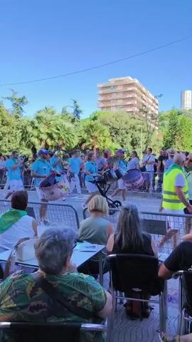 The video shows an outdoor event with a brass band performing on a street. The band members are wearing matching light blue shirts and white shorts, and they are playing various instruments including drums, trumpets, and saxophones. They are positioned behind a metal barricade, with spectators seated at tables in the foreground. In the background, there are trees and a multi-story building. The sky is clear and blue, indicating it is a sunny day.
