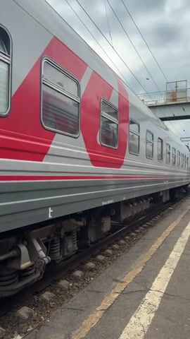 This is a side view of a grey train car with red markings on the side. The train is stopped at a station, with tracks visible in the foreground. The sky is overcast. There are no people visible in the frame.