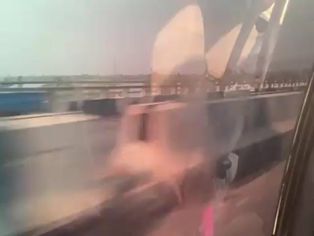 A car is driving next to a flatbed truck carrying metal beams. The car passenger is filming the truck from the side window. The flatbed truck passes the car. The view shows the driver and the car interior, including a rear view mirror. The landscape is visible in the distance.