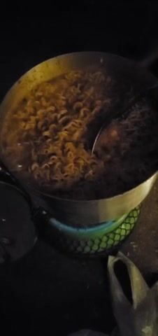 A video shows a large pot of noodles cooking on a small portable stove with a green mesh exterior. The pot appears to be stainless steel and contains a large quantity of cooked noodles in a broth. A ladle rests inside the pot, partially submerged in the noodles. A small flame is visible beneath the pot, indicating the stove is lit. The scene is dimly lit, with only the glow from the stove and pot illuminating the area. A dark, possibly metal, object is visible to the left of the stove, partially out of frame. A white plastic bag is visible at the bottom right corner of the frame. The setting appears to be outdoors or in a poorly lit area.
