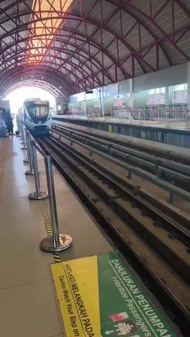 The video shows a light rail train pulling into a covered station. The train is white and blue with its headlights on and a green light illuminated on the front. It is traveling on parallel tracks from left to right in the frame. A platform is visible on the left side of the tracks, separated from the tracks by a yellow and silver barrier. There are several people on the platform. A yellow and green sign is visible on the platform with text in both Malay and English, indicating to watch your step and prioritize passengers. The station has a distinctive arched roof structure with red framing and metal siding. The lighting suggests it is daytime, with sunlight visible in the distance at the end of the tunnel where the train is entering. The overall environment appears to be a modern public transportation station.