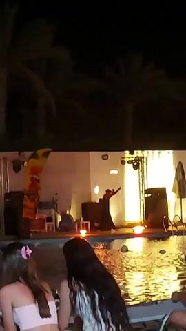 At night, in front of a pool, a performer is doing a fire show. The performer is in a dark outfit and is spinning a flaming object. The flames are visible in the water of the pool and in the air. There are palm trees in the background. Two young women are sitting by the pool, watching the performance. They appear to be enjoying themselves.