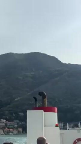 The view from a boat shows two rusty pipes emitting smoke from a structure with a red and white top. A forested mountain range stands in the background, with a few houses visible on the lower slopes. A sliver of water can be seen at the bottom of the frame. A man's bald head is briefly visible at the bottom of the frame.