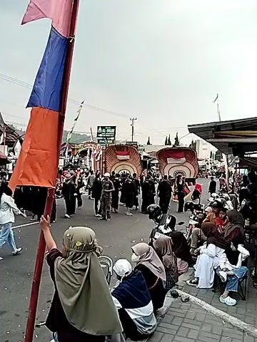 The video shows an outdoor event, likely a parade or festival. In the foreground, on the right side of the street, several people are sitting on chairs and the curb, observing the procession. They are mostly wearing casual clothing, with some individuals wearing hijabs. A motorcycle is parked in front of them. On the left side of the street, a person is holding a large flag pole with red, white, and blue flags. In the middle of the street, a group of people in black clothing are walking. They are escorting two large, elaborate floats that resemble giant bird heads or masks, decorated with the Indonesian flag. Several spectators are lining the sides of the street, watching the procession. In the background, there are buildings, signs, and trees. The sky is overcast. The overall impression is a lively community gathering.