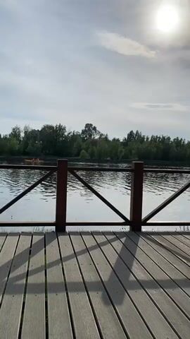 The video pans across a wooden boardwalk overlooking a calm body of water, likely a lake or river. The sun shines brightly in a cloudy sky, casting shadows on the wooden planks. On the water, a small yellow kayak with two people can be seen in the distance. The opposite bank of the water is lined with lush green trees. In the background, several modern buildings are visible, one with a distinctive domed roof. The video captures a serene outdoor scene on a partly cloudy day.
