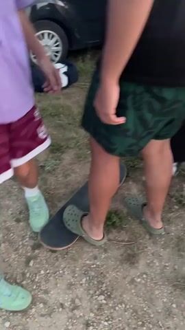 The video shows a group of young people outdoors, possibly at dusk. The focus is on a person in the foreground wearing a black t-shirt and green patterned shorts, standing with one foot on a skateboard. Another person in a purple and white shirt is partially visible to the left, also near the skateboard. In the background, several cars are parked along a dirt road, and a few more people are visible. The lighting suggests it is late afternoon or early evening. The scene appears casual and relaxed.