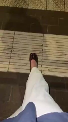 The video shows a person wearing a dark, shiny shoe with a split toe design. The shoe is worn on their right foot. The person is also wearing white pants. The video focuses on the shoe and foot, showing close-up views from the top and side. The person moves their foot slightly. The background is a set of stairs with light-colored, horizontal treads.