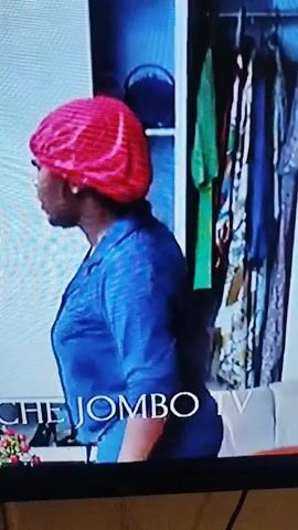 A medium shot captures a woman in a blue outfit and a red hair bonnet standing in a room. The room features a couch with a red pillow, a window with grey curtains, and a closet with hanging clothes. The woman is turned to her right and appears to be yelling or speaking intensely. The words "UCHE JOMBO TV" are superimposed on the bottom of the screen.
