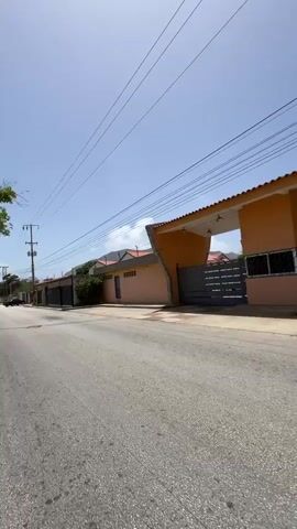 The video captures a motorcycle journey on a sunny day in Los Cerritos, Venezuela.

The initial shot (0:00-0:02) shows the motorcycle approaching a peach-colored gated entrance to what appears to be a residential complex. The gate features two large arched sections with a small central building, and dark metal gates. Overhead, multiple power lines crisscross against a clear blue sky.

As the motorcycle passes the gate (0:02-0:05), the view shifts to a long, neatly trimmed green hedge lining the side of the road, with buildings visible beyond it. The buildings are light-colored, some with multiple windows, and topped with terracotta-style roofs. A concrete wall with a security fence on top is also visible.

From 0:05 to 0:08, the perspective shifts to a first-person view behind a mint green motorcycle helmet. The helmet has "Ridery" and "Rider" branding, along with a sticker displaying additional "Rider" and "Ridery" text, and what appears to be a phone number ("0412-296.301") and another number ("0865319"). The rider's light blue shirt is also visible. Another motorcycle is seen ahead on the paved road, which appears to be curving gently to the right. The sky remains clear and bright, and power lines are still visible overhead.