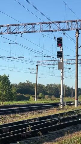 The video shows a railway track with overhead power lines and a signal light displaying a red signal. The signal light is mounted on a tall pole, and there are multiple sets of tracks visible in the foreground. Trees and foliage can be seen in the background under a clear blue sky. The scene appears to be set during the daytime.
