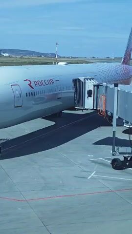 An airplane with the word "ROSSIA" written on its side is parked at an airport gate. A jet bridge is connected to the airplane. Two people are visible near the front of the plane. The sky is blue with some clouds. The ground is paved with asphalt and marked with yellow and white lines.
