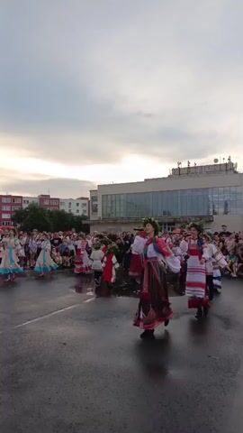This media file depicts an outdoor cultural event, likely a festival or performance, taking place in Kryvdanivka, Russia. The scene is set on an asphalt ground, possibly a public square or a performance area, with a crowd of spectators gathered in the background. Behind the crowd, a modern building with large glass windows and a distinctive architectural style is visible, suggesting an urban or semi-urban setting. Residential buildings with pink and white facades are also present in the distance.

The foreground is dominated by a group of performers, mostly women and girls, dressed in vibrant, traditional folk costumes. These costumes are characterized by intricate embroidery, colorful patterns, and flowing skirts, some adorned with wreaths of flowers on their heads. They are engaged in a lively dance or choreographed movement, their colorful attire creating a dynamic visual spectacle. Some male performers in similar traditional attire are also present. The performers appear energetic and joyful, their movements suggesting a celebratory atmosphere.

The time of day appears to be late afternoon or early evening, with the sky filled with dramatic, cloudy formations, indicating a possibly overcast or approaching weather change. The ground is wet and reflective, suggesting that it may have recently rained or that the area is prone to puddles, which adds an interesting textural element to the scene.

The overall mood conveyed is one of cultural celebration and community gathering, with the bright costumes and energetic performances contrasting with the muted tones of the sky and the surrounding buildings. There is no visible text in the media file.