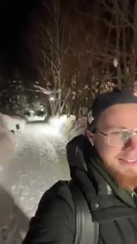 A man with a red beard and glasses is walking along a snow-covered path at night. He is wearing a black jacket, a navy cap, and white earphones. The path is lined with trees and illuminated by streetlights. The man is smiling and looking at the camera. The video appears to be a selfie video.