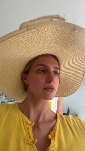 A woman in a yellow shirt and a very large straw hat puts on heart-shaped pink sunglasses. She is in a store, and there is music playing in the background. Another person is visible standing behind her. The woman looks at the camera while adjusting the sunglasses.