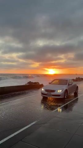 A silver coupe is parked on a wet, paved road next to a stone seawall. The ocean is to the left of the car, with waves crashing against the seawall. The sky is filled with clouds, and the sun is setting, casting an orange glow on the clouds and the water. The car's headlights are on, and their reflections can be seen in the wet pavement. There is a distant pier or structure in the ocean.
