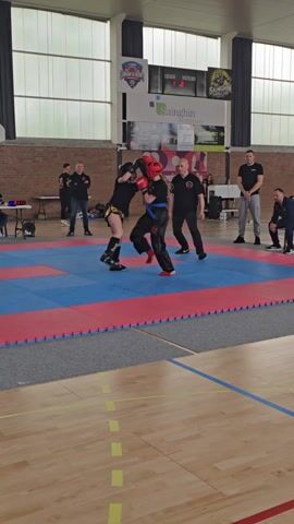 Here is a description of the video:

Two young people wearing headgear, gloves, and protective clothing are sparring on a blue and red padded mat. They are in a gymnasium or large indoor sports facility with a hardwood floor marked with yellow lines. Several adults are watching the match, including two in black shirts who appear to be officiating or coaching.  The background includes a brick wall, windows, and banners displaying logos and text in French. A scoreboard indicates scores for 'Locaux' and 'Visiteurs'.  The camera is static and captures a wide view of the sparring area.
