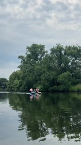Two individuals are seen on a body of water, each on a paddleboard. The person in front appears to be wearing a pink shirt, while the person behind them is wearing a darker top. They are paddling on a calm river or lake, with lush green trees lining the bank in the background. The sky is cloudy.
