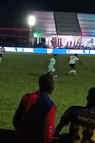 The video shows a soccer game being played at night. The field is illuminated by stadium lights and the backdrop includes a large tent or structure adorned with colorful lights and banners. In the foreground, three people are watching the game, partially obscuring the view. On the field, several players are actively engaged in the game, dressed in contrasting uniforms. The players are running and maneuvering the ball, indicating a dynamic play in progress. The overall environment suggests a lively and festive atmosphere surrounding the soccer game.