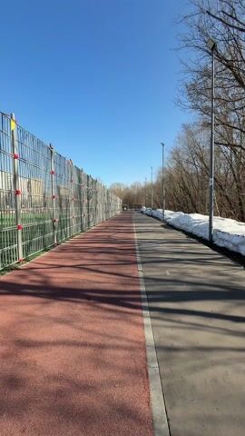 A woman takes a selfie video while walking on a red and gray path outdoors on a sunny day. The path is bordered by a tall chain link fence on one side, and trees and patches of snow on the other. Street lights are visible along the path. The woman is wearing a black hoodie and has her hair pulled back in a ponytail. She is smiling and looking at the camera. A person can be seen running in the distance.